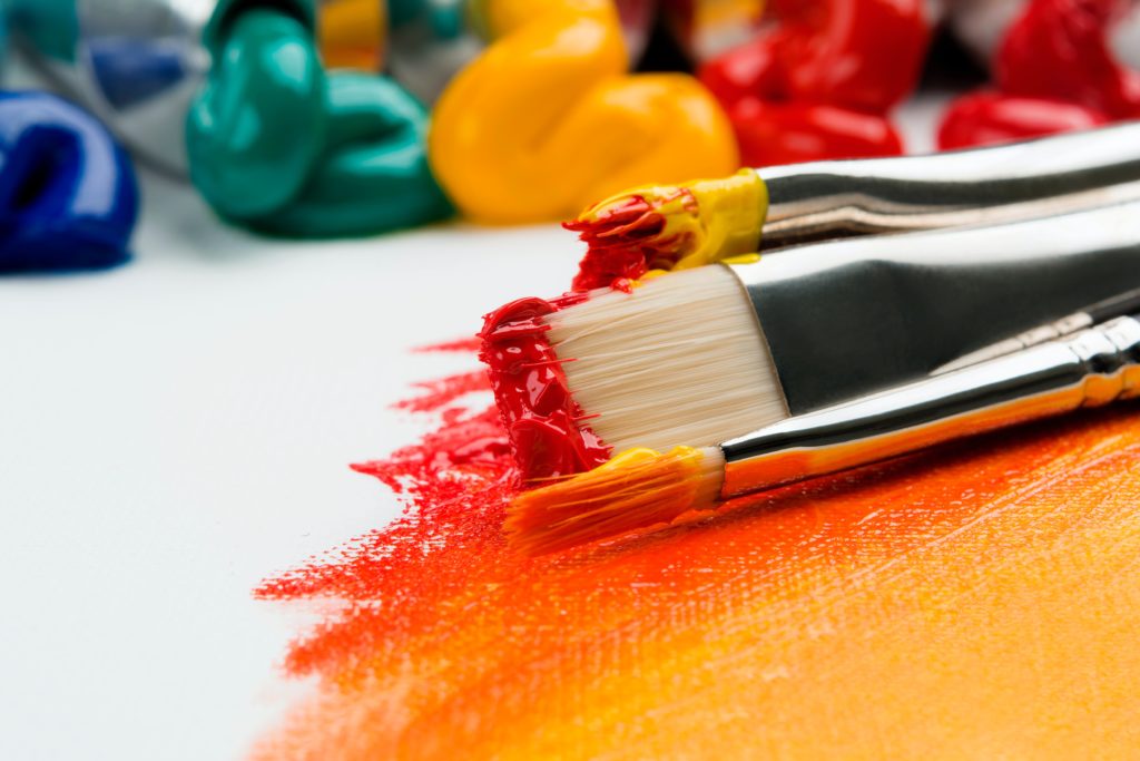 paint brushes and paints laying on a canvas, showcasing reds and yellows with green and blue paint coming out of tubes in background