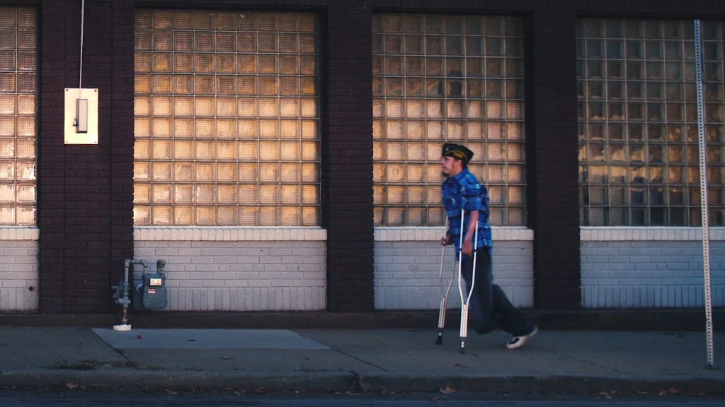 VFW, Veterans, Sacrifice, Sgt. Scott Stephenson - a video production by scenic road - man walking down the street along a subway block glass backdrop on crutches.