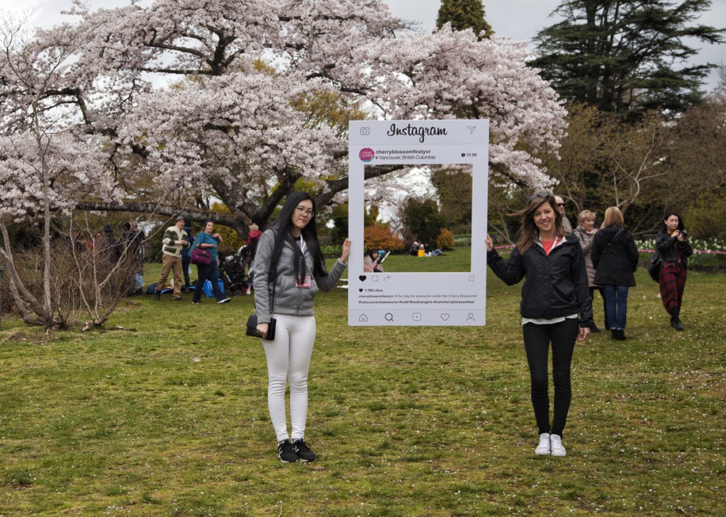 Two girls holding a picture frame of instagram in front of a blossoming cherry tree.