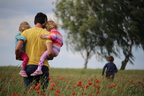 man-in-yellow-shirt-carrying-two-daughters-through-field-of-poppies-while-boy-runs-ahead