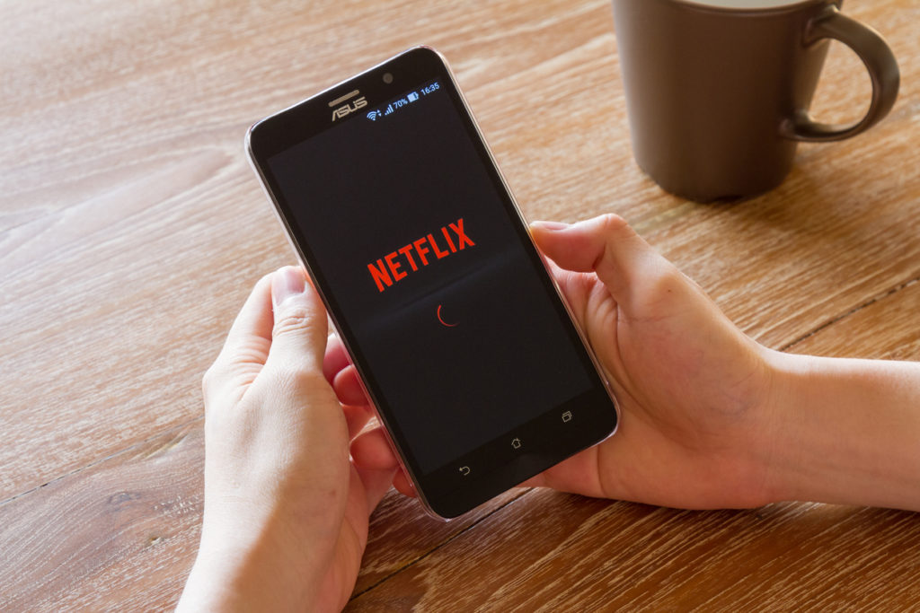 hands holding mobile device showing Netflix app on top of wooden table